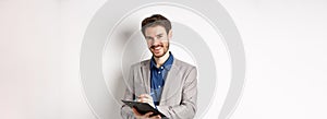 Smiling male manager in suit writing on clipboard, taking notes at office meeting, standing on white background