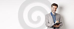 Smiling male manager in suit writing on clipboard, taking notes at office meeting, standing on white background