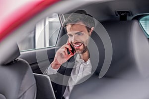 Smiling male sitting at backseat of car with laptop, talking on phone