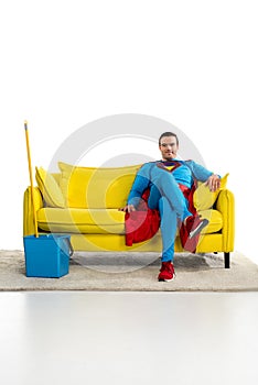 smiling male superhero sitting on couch near bucket and mop
