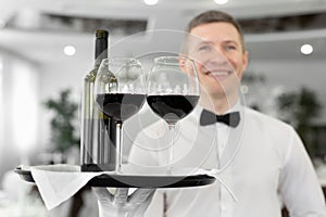 Smiling male waiter with glasses of red wine and a bottle on a tray in a restaurant