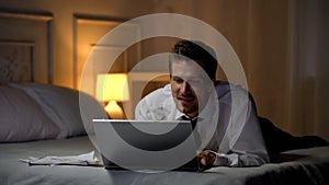 Smiling male in white shirt on bed smiling and chatting on laptop, office affair