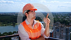 Smiling male worker in helmet counting and pointing with a finger at the construction site on the roof. Business