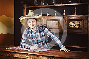 Smiling man as bartender in a sombrero leaned on bar counter in