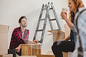 Smiling man assembling a cabinet and his girlfriend drinking coffee while furnishing interior