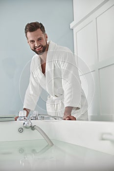 Smiling man in bathrobe looks at modern hydro massage tub full of hot water in salon