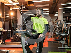 A smiling man with a beard rides a bike in the gym, listens to music and takes selfie