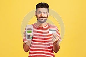 Smiling man with beard in striped t-shirt holding and showing pos payment terminal and credit or debit card, using cashless