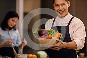 A Smiling Man with the Bowl of Fresh Vegetables, Preparing for Vegan Food in a Modern Kitchen. Blurred Woman Stirring a Healthy