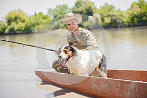 Smiling man in camouflage and hat fishing with setterdog, while wading in muddy river on old boat.