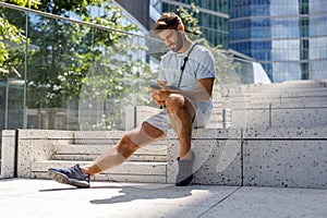 Smiling man in casual clothes sitting on skyscrapers background and listen music in headphones