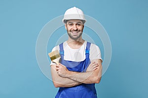 Smiling man in coveralls protective helmet hardhat hold paint brush isolated on blue wall background. Instruments