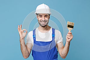 Smiling man in coveralls protective helmet hardhat hold paint brush isolated on blue wall background. Instruments