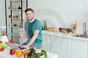 smiling man with credit card sitting at table with laptop in kitchen