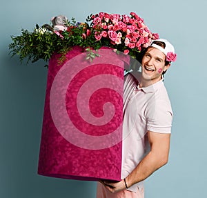 Smiling man delivery guy with rose at ear and lipstick kiss on his cheek is holding huge valentines day box with flowers