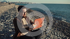 Smiling man is doing video call by tablet on a pebble beach, waving hand