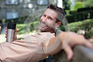 smiling man drinks coffee sitting on city bench