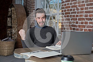 Smiling man freelancer raising hand in yes gesture, celebration victory, looking on laptop computer.