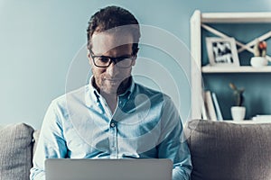 Smiling Man in Glasses with Laptop on Gray Sofa.