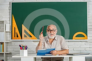 Smiling man in glasses sit at desk. Old man teachers on green board. Happy school male teacher book. Portrait male