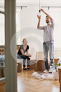 Smiling man hanging lamp while furnishing new home after moving in with his happy wife