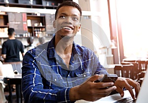 Smiling man holding cellphone with laptop in cafe