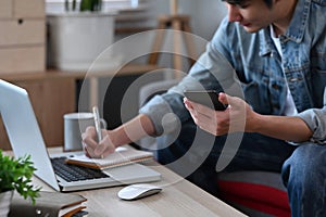 Smiling man holding mobile phone and making notes on notebook while working from home.