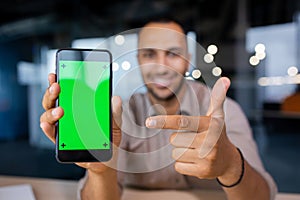 Smiling man holding a phone with a green screen in his hands, showing it to the camera and pointing at it with his index