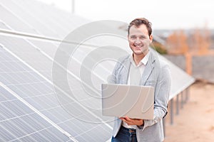 A smiling man, holds an laptop, standing next to the solar panel. Outdoors