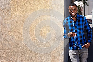 Smiling man leaning against wall with smart phone and earphones