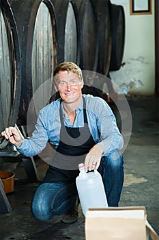 Smiling man middle-aged working in winery pouring wine from wood to plastic bottle in cellar