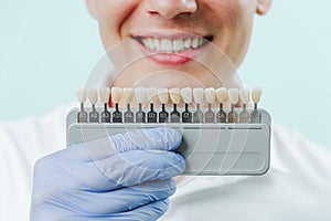 Smiling man mouth with natural white teeth in light blue background in dental clinic. Hands holding the teeth color palette next