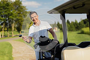 A smiling man pulls out of a bag with sticks, a golf club. Bag lies on the luggage compartment of the golf cart