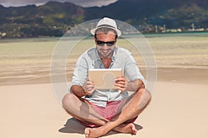 Smiling man reading on tablet while sitting down the beach