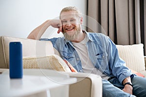 Smiling man relax on sofa look at camera use portable wireless speaker on table