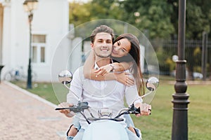 Smiling man with short hairstyle driving scooter down the green street while his long-haired girlfriend huggs him