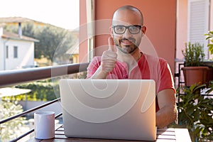 Smiling Man sitting at his wooden desk working on laptop showing thumb up. Boy using computer. Positive caucasian man in red t-shi