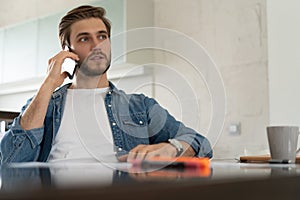 Smiling man, he is sitting at a table, talking on the phone with his laptop, notebook in front of him