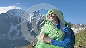 Smiling man in a sleeping bag with a thermo mug in his hand looks at the sun at dawn in the mountains. The hiker woke up