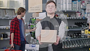 Smiling man stretching box at camera with blurred customer choosing tools at background. Portrait of positive Caucasian