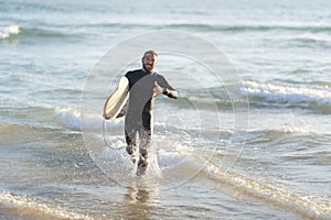Smiling man surfer running out of the sea holding a surfboard and showing shaka