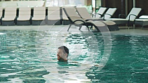 Smiling man swimming at modern hotel. Sexy guy bathing at luxury pool indoor.