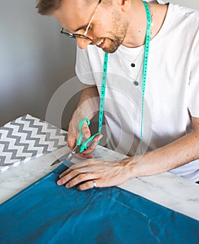 A smiling man tailor cuts fabric in a home studio. A self-taught seamster in white t-shirt and glasses works with fabric, pattern