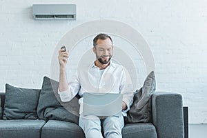 smiling man turning on air conditioner with remote control while using laptop