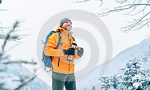 Smiling Man under snowfall dressed bright orange softshell jacket with a hot drink thermos flask while he trekking winter