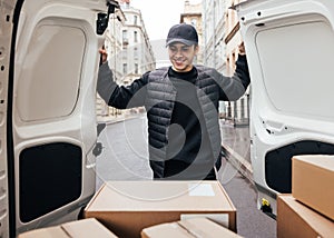 Smiling man in uniform and cap looking at cardboard boxes while standing at van trunk