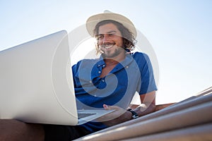 Smiling man using laptop while relaxing on hammock at beach