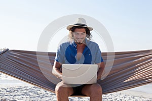 Smiling Man using laptop while sitting on hammock at beach