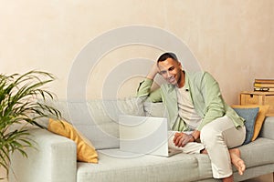 Smiling Man Using Laptop At Sofa. Guy Relaxing With Laptop At Home On Cozy Couch