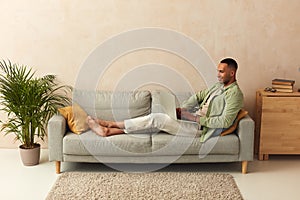 Smiling Man Using Laptop At Sofa. Guy Relaxing With Laptop At Home On Cozy Couch